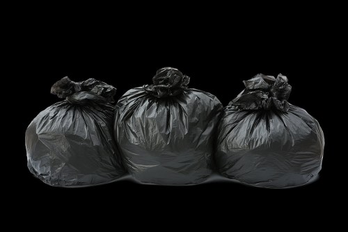 Three large black rubbish bags made of thick plastic, tightly knotted at the top, are arranged on a dark background. The bags appear full and slightly crumpled, with visible creases and uneven surfaces indicating they are filled with waste. The bags are positioned side by side, with the middle bag slightly leaning forward, and the surrounding area is black, giving a clear focus on the waste containers. This image likely relates to a rubbish removal service, such as those offered by Garden Clearance Clapham, suitable for garden or household waste disposal in the Clapham area. The plastic material has a matte finish, and the lighting highlights the texture and contours of each bag, emphasizing their bulging form and the tightly gathered tops. The clean, dark background isolates the bags, presenting a straightforward visual of typical waste bags awaiting collection or disposal, relevant to rubbish collection and clearance services in the local London postcode areas.