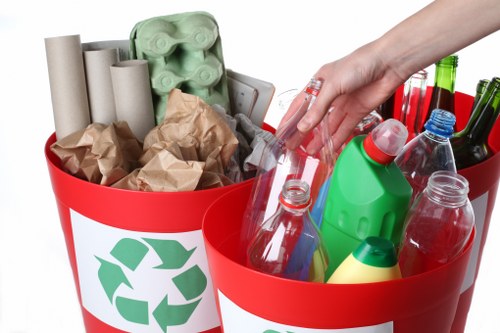 A close-up of two red recycling bins with white recycling symbols, placed outdoors on a driveway or pavement. The bin on the left contains crumpled brown paper, several empty cardboard tubes, and a green plastic egg carton. The bin on the right holds various empty plastic bottles of different shapes and sizes, including clear, green, and blue bottles, some with caps still attached. A hand with a fair complexion and neatly trimmed nails is reaching into the bin on the right, as if about to add or remove an item. The scene is well-lit with natural daylight, and the background is slightly blurred, suggesting an outside setting typical of rubbish collection or garden clearance activities in Clapham or nearby areas. The context aligns with waste disposal or recycling services offered by Garden Clearance Clapham, focusing on rubbish removal from domestic or garden environments.