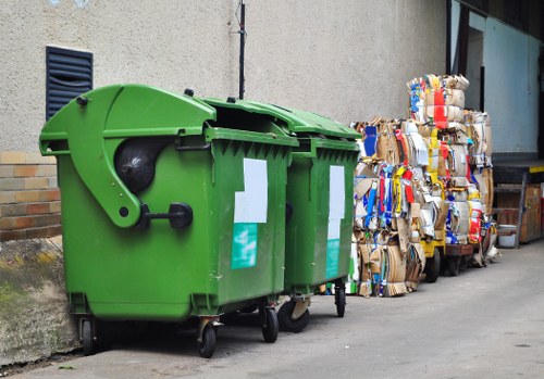 A bright green two-compartment wheelie bin made of durable plastic with a textured surface, positioned on a paved area beside a light grey exterior wall of a building. The bin has black wheels at the base for mobility, a black handle, and a closed lid with small ventilation holes on the side. Next to the bin, several tightly packed bales of flattened cardboard and paper waste are stacked, showing multi-colored cardboard packaging and various shades of brown and grey paper, indicating nearby rubbish removal activity. The scene appears to be outdoors, possibly on a residential or commercial pavement, with a portion of a window or glass door with a dark frame visible in the background, suggesting proximity to a property in Clapham or surrounding London suburbs. The environment is well-lit, with natural daylight illuminating the objects and surroundings, highlighting the clean, organized storage of waste materials for collection by a waste management service like Garden Clearance Clapham, specializing in rubbish removal.