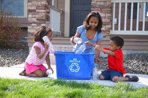 Three children, two girls and one boy, are sitting on a light-colored outdoor mat on a well-maintained grassy front garden near a residential property, actively filling a blue recycling bin with clear plastic bottles. The recycling bin, positioned centrally in the image, features a white recycling symbol and the words 'WE RECYCLE' printed on it, indicating a focus on environmentally friendly waste management. The children are engaged in the collection activity: the girl on the left, wearing a pink and white striped top and pink skirt, is holding a plastic bottle close to her mouth; the girl in the middle, dressed in a light blue top, is pouring a plastic bottle from her hand into the bin; and the boy on the right, wearing a red t-shirt and dark shorts, is also placing a bottle into the bin with his right hand. The background shows a residential building with stone and white-painted wood siding, a front door, and a porch area with white railing. The garden area features neatly trimmed grass, decorative gravel, and some small bushes, suggesting a tidy outdoor space suitable for recycling or rubbish collection activities managed by Garden Clearance Clapham.