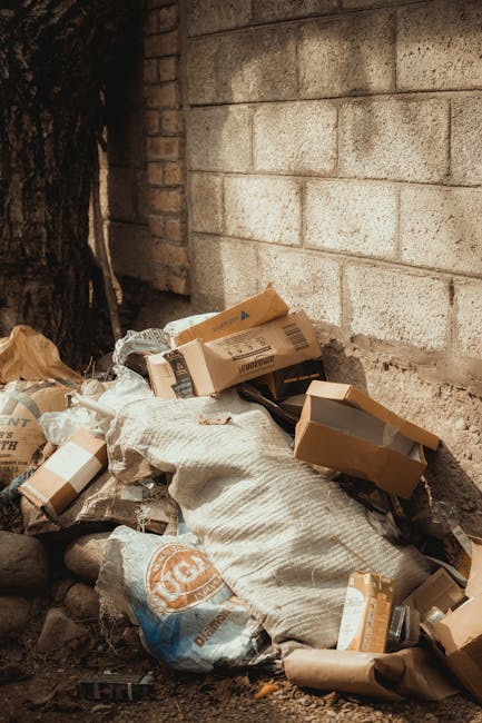 An outdoor scene showing a pile of discarded waste materials positioned against a rough brick wall and a concrete block wall. The pile includes flattened cardboard boxes of various sizes, some with vi