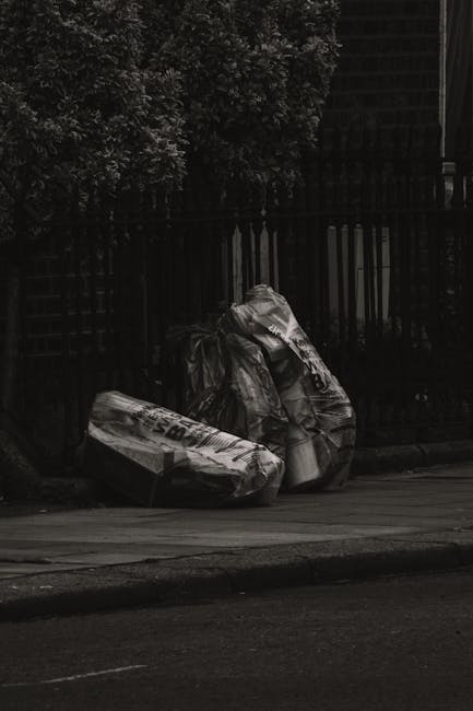An overflowing collection of waste and rubbish bags surrounding multiple black, grey, and red wheelie bins on a paved urban street. The rubbish includes a mix of cardboard boxes, paper, plastic bags, 