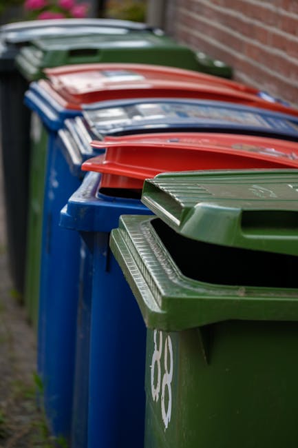 Two large black plastic garbage bags filled with rubbish are placed on the sidewalk in front of a black metal fence and behind a set of leafy green trees. The bags appear to contain household waste or