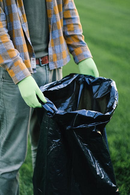 Three large black rubbish bags made of heavy plastic material are stacked against a black metal fence on a paved sidewalk. One bag appears to be kinked and partially collapsed, while the other two are